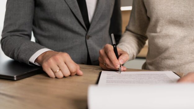 Contract ondertekening aansprakelijkheid Twee personen ondertekenen een contract aan een bureau, passend bij het thema aansprakelijkheidsuitsluiting.