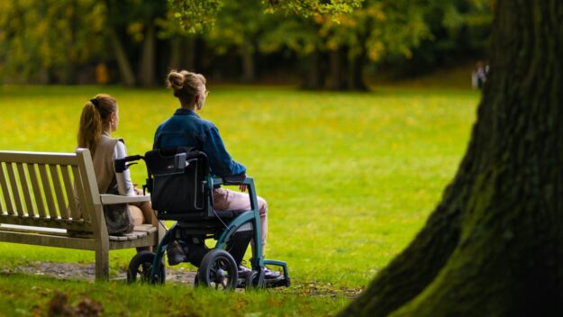 Twee vrouwen in gesprek, één op een bankje en één in een rolstoel, kijkend naar een groen park