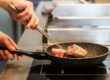 Een hand draait een stuk vlees om in een pan op een inductiekookplaat, tijdens het inductie koken.