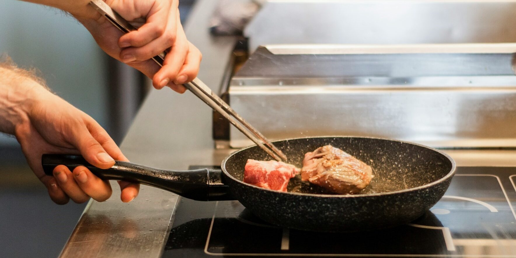 Een hand draait een stuk vlees om in een pan op een inductiekookplaat, tijdens het inductie koken.