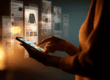 Persoon met smartphone, omringd door licht van het scherm en online reclamebeelden. De foto laat zien hoe digitaal consumentisme ons dagelijks leven beïnvloedt.
