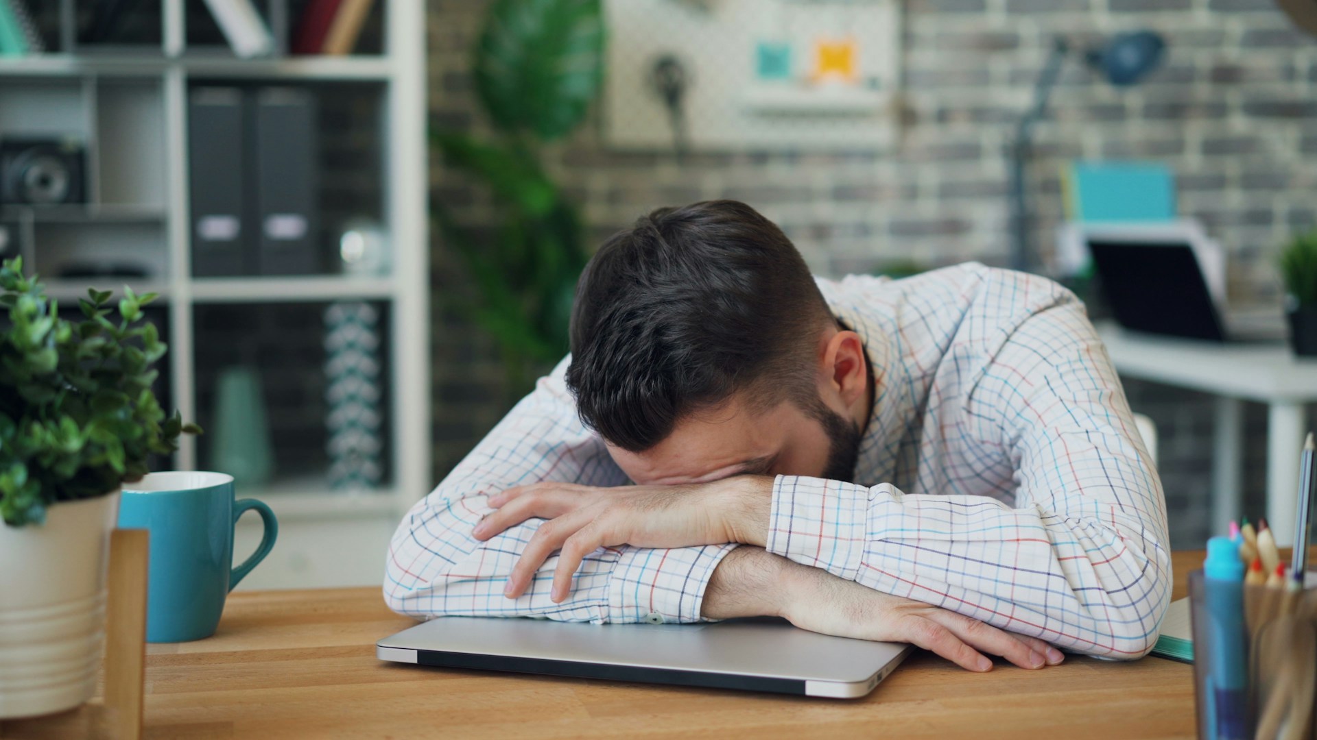 Man ligt uitgeput met hoofd op armen naast laptop als gevolg van langdurige prestatiedruk